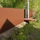 Забор из коричневого профнастила и мостик, перекинутый через траншею.